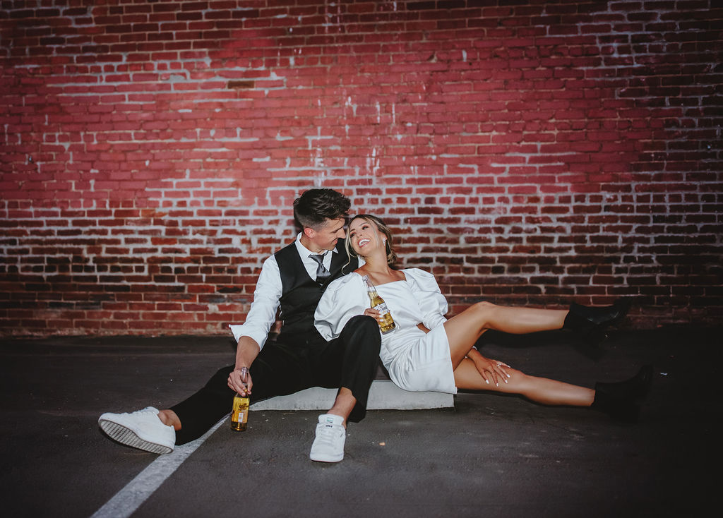 Bride and groom drinking coronas in front of brick wall in Fresno, CA