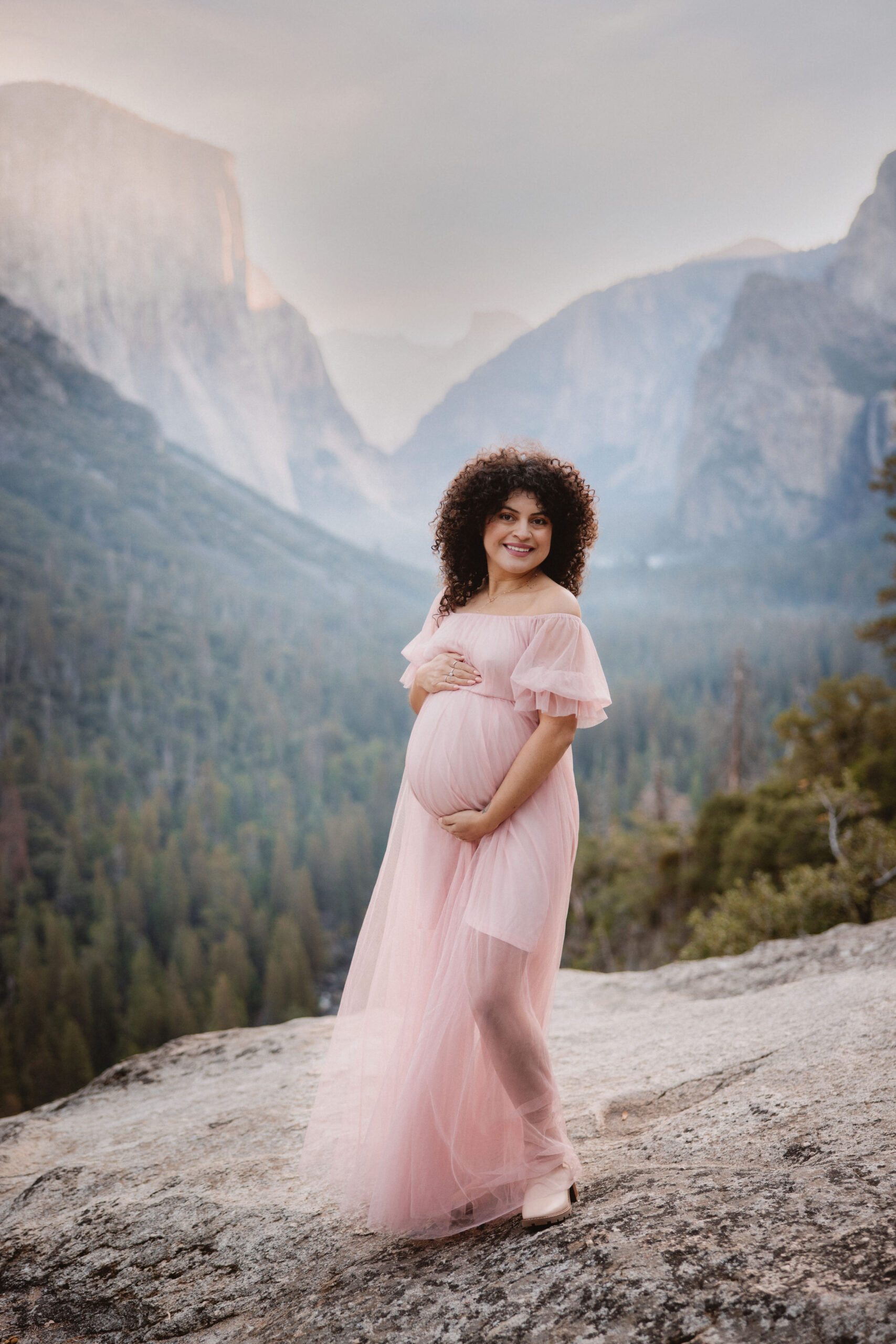 Pregnant woman in a pink dress standing on a rocky terrain with a scenic forest and mountain backdrop during sunset at her maternity photoshoot