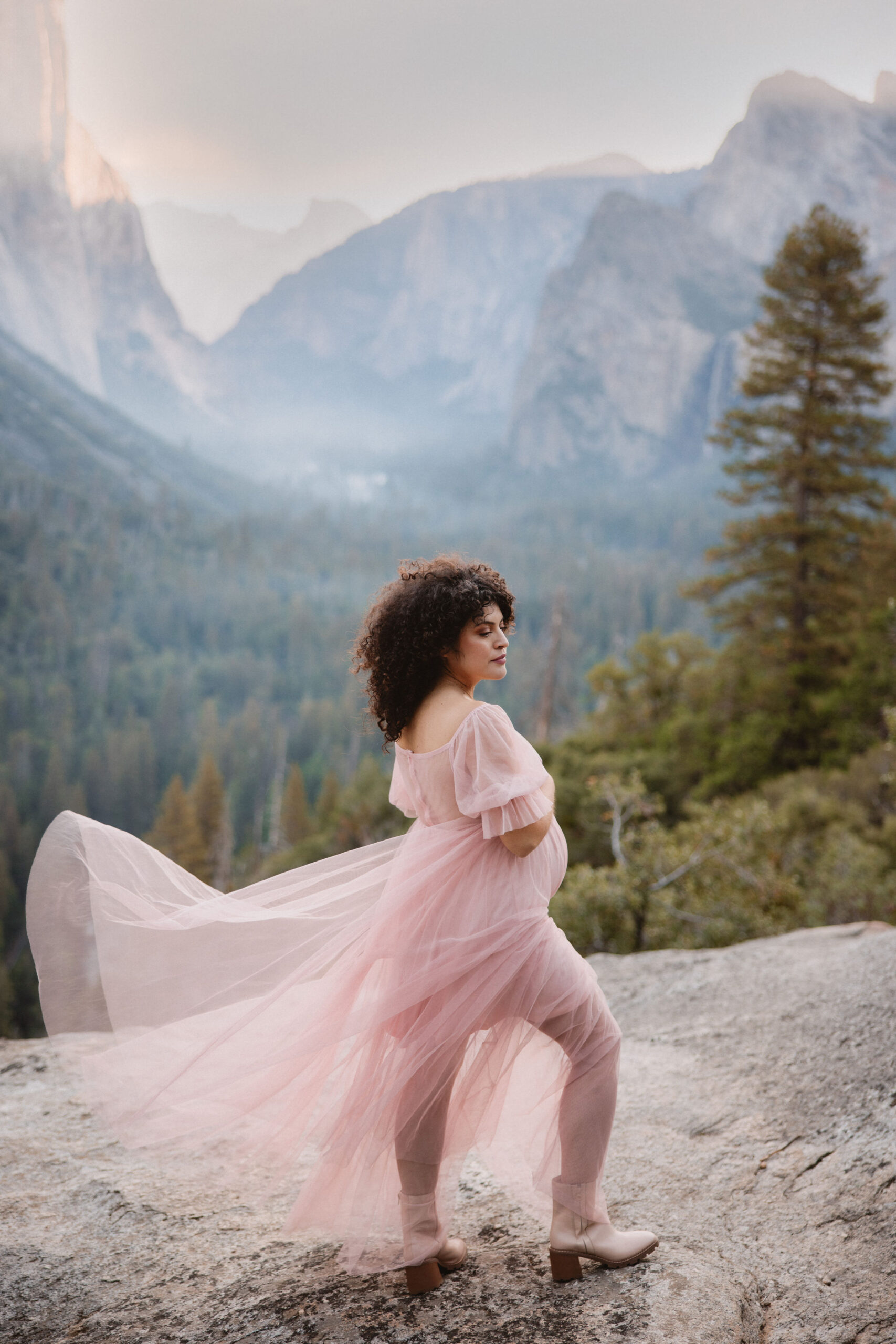 Pregnant woman in a pink dress standing on a rocky terrain with a scenic forest and mountain backdrop during sunset at her 