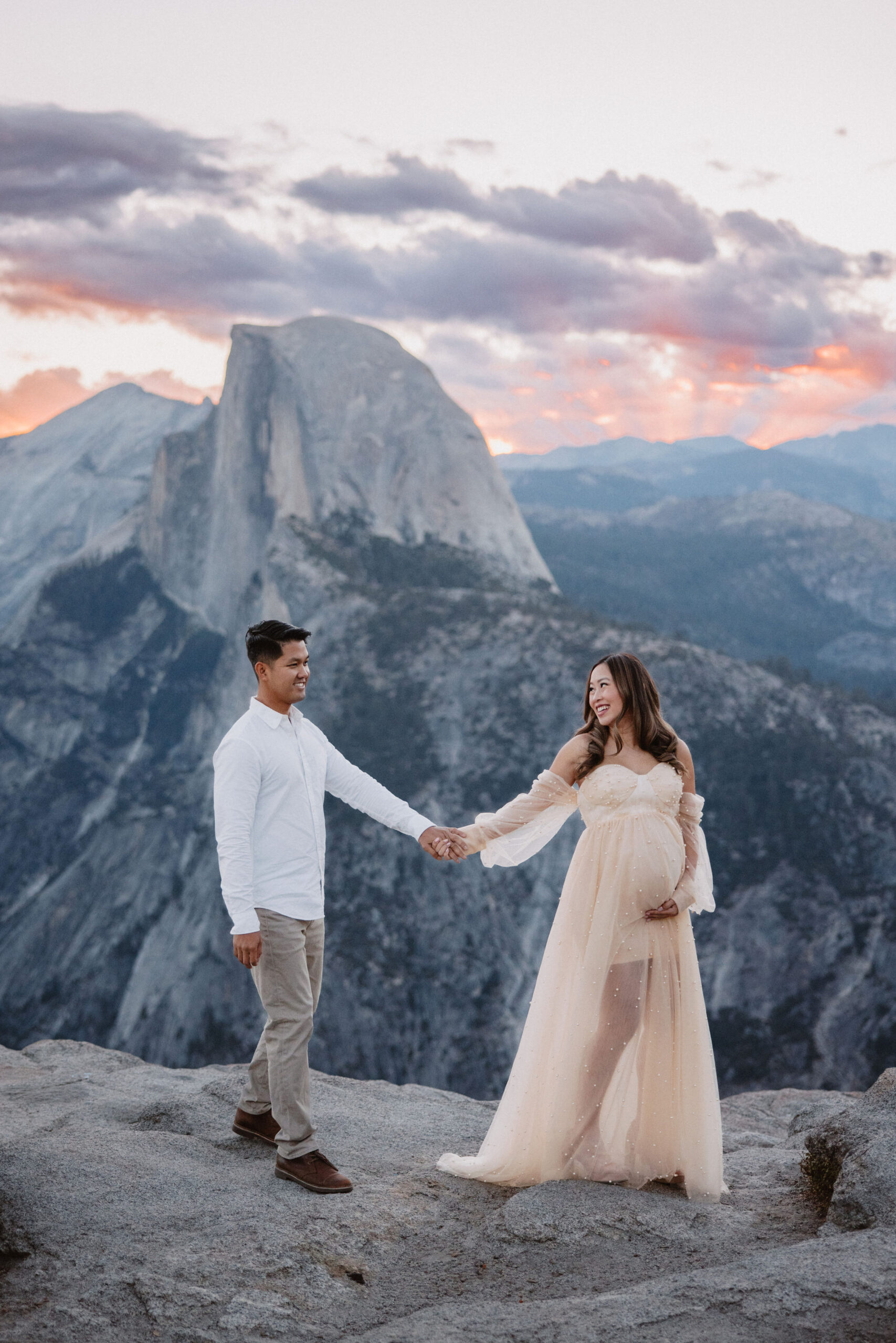 A pregnant woman in a beige dress stands thoughtfully on a rocky overlook, with yosemite valley's iconic cliffs and forests in the background at her maternity photoshoot