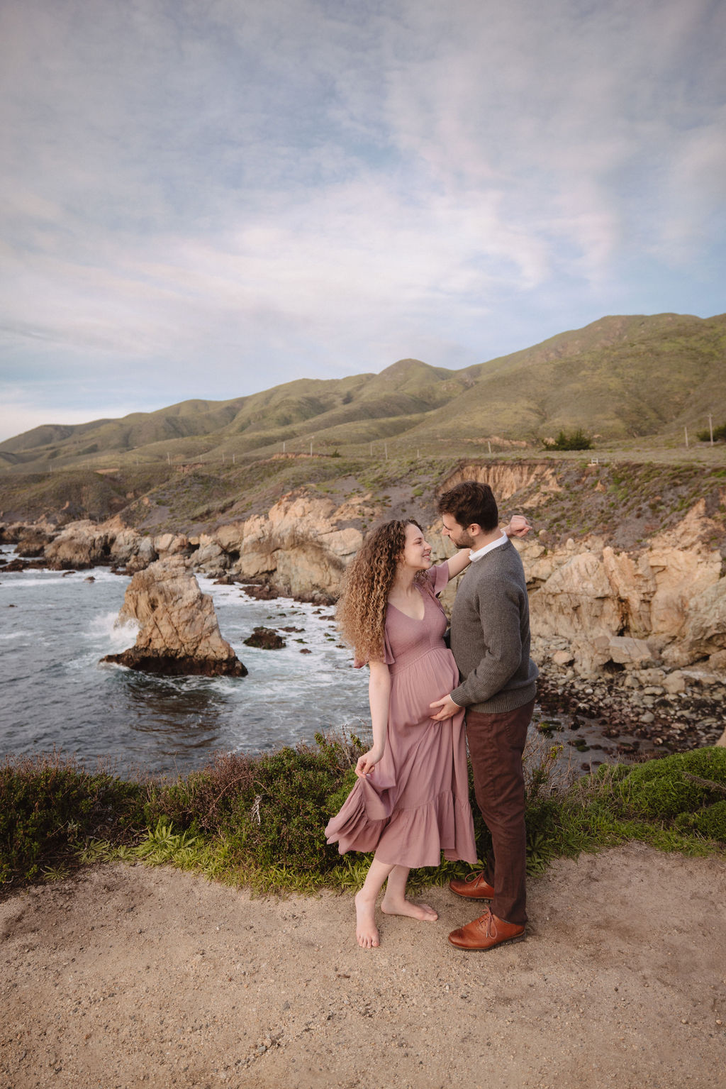 A couple embracing and smiling at each other on a scenic coastline with hills in the background at their beach engagement photos. 