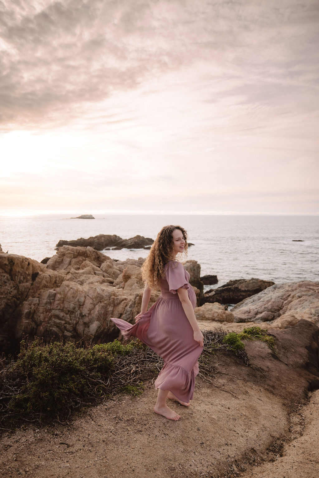 Pregnant woman in a pink dress standing by the seaside with hills in the background to capture her beach maternity photos