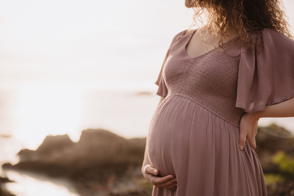 Pregnant woman in a pink dress standing by the seaside with hills in the background to capture her beach maternity photos