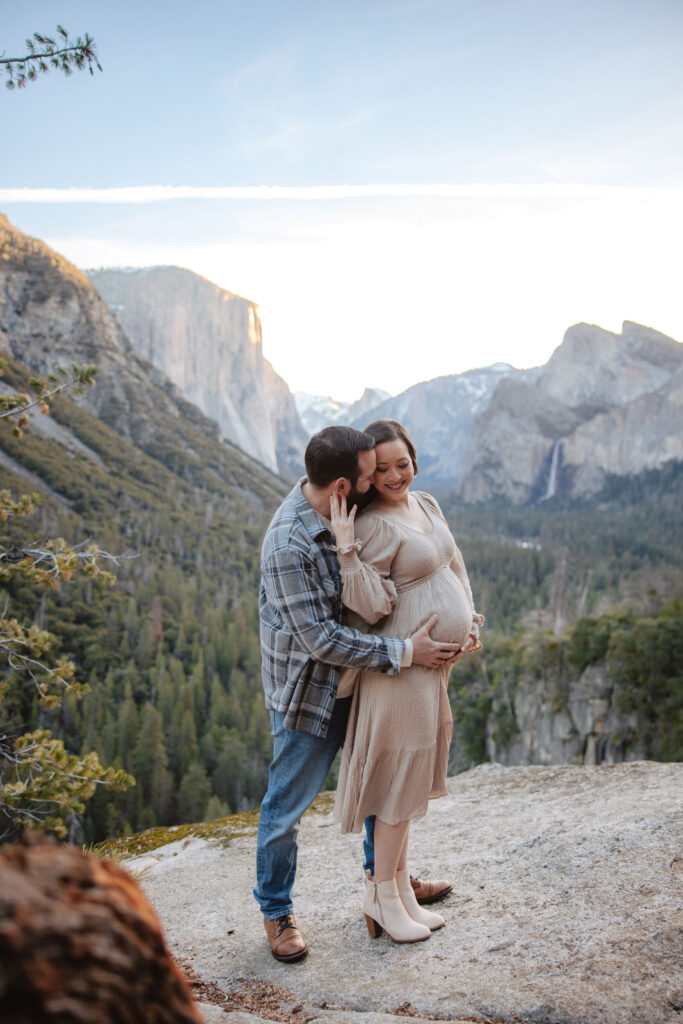 A couple embraces on a mountain overlook at sunset, with a pregnant woman smiling as her partner kisses her forehead, scenic cliffs and forests in the background at her maternity photoshoot