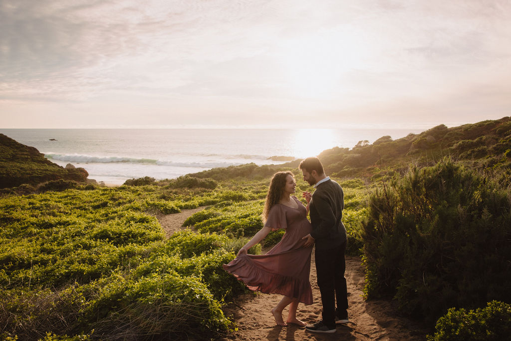 A couple embracing and smiling at each other on a scenic coastline with hills in the background at their beach engagement photos. 