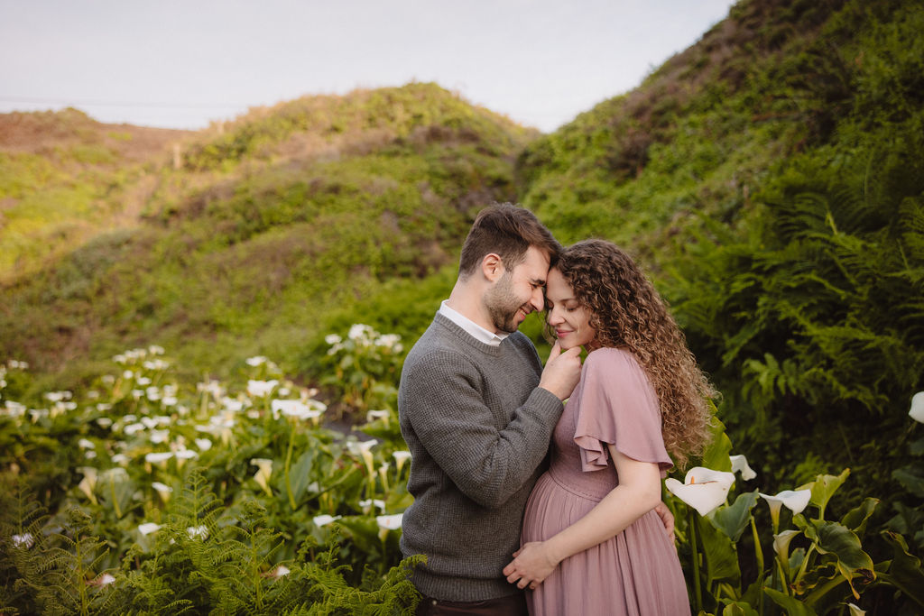 A couple expecting a child, sharing an intimate moment outdoors among calla lilies at their beach engagement photos