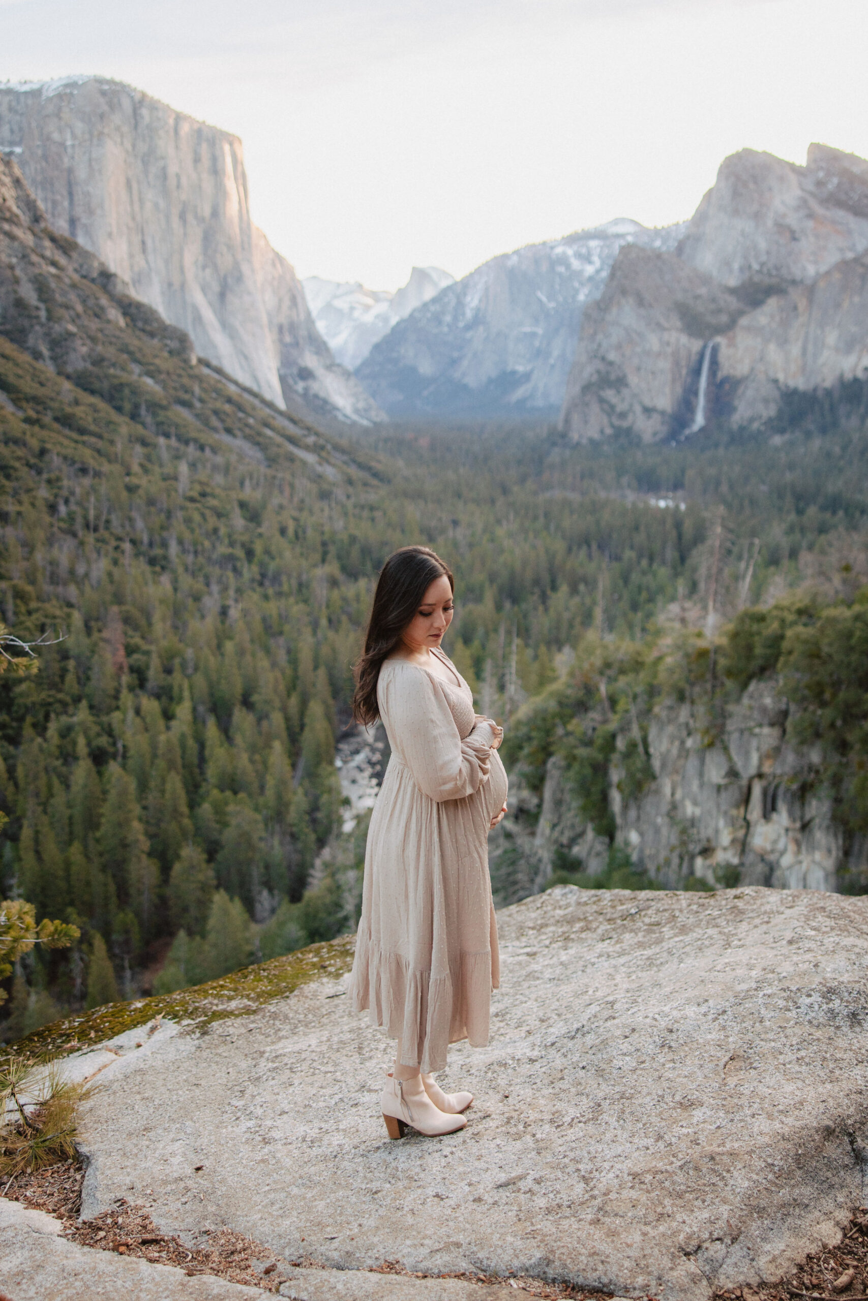 A pregnant woman in a beige dress stands thoughtfully on a rocky overlook, with yosemite valley's iconic cliffs and forests in the background at her maternity photoshoot