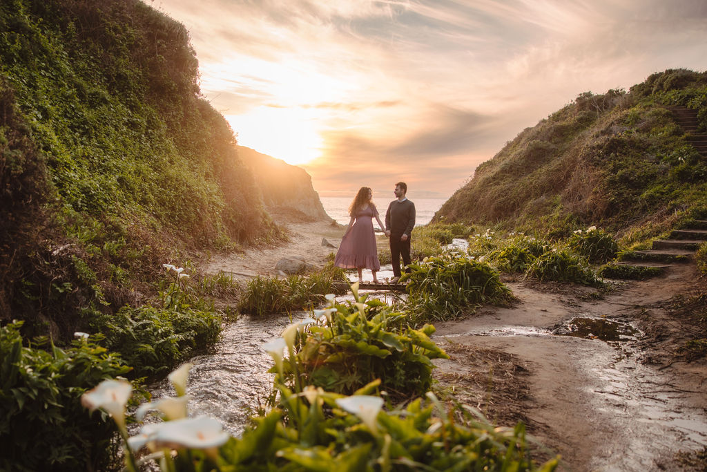 A couple holding hands and walking towards the beach at sunset, with flowers and a pathway in the foreground at Big Sur