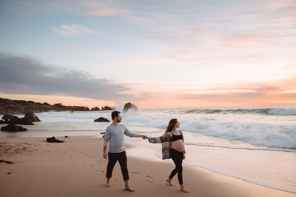 A couple holding hands and walking on a beach at sunset during their beach maternity photos