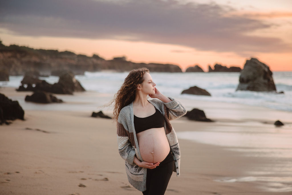 Pregnant woman standing on the beach at sunset, cradling her belly.