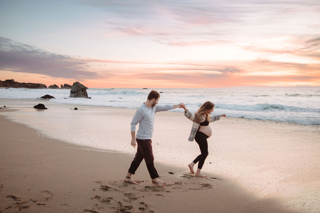 A couple playfully dancing on a sandy beach at sunset, with waves lightly brushing the shore and rocks in the background.