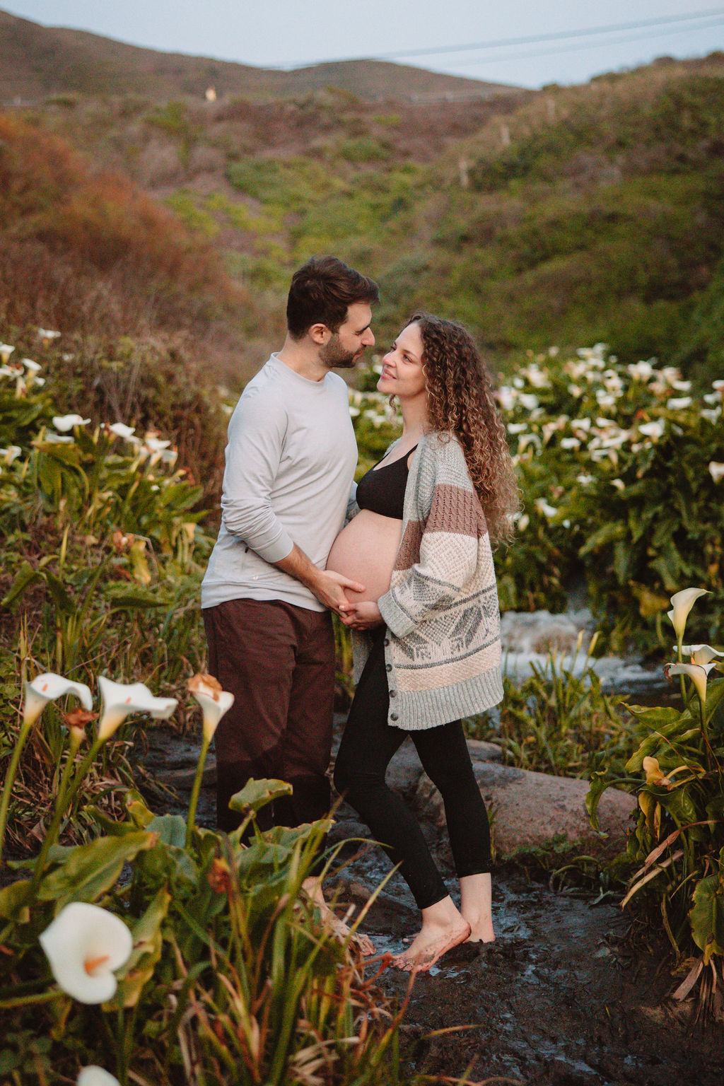 A couple expecting a baby, sharing a tender moment amidst a field of white flowers at Big Sur