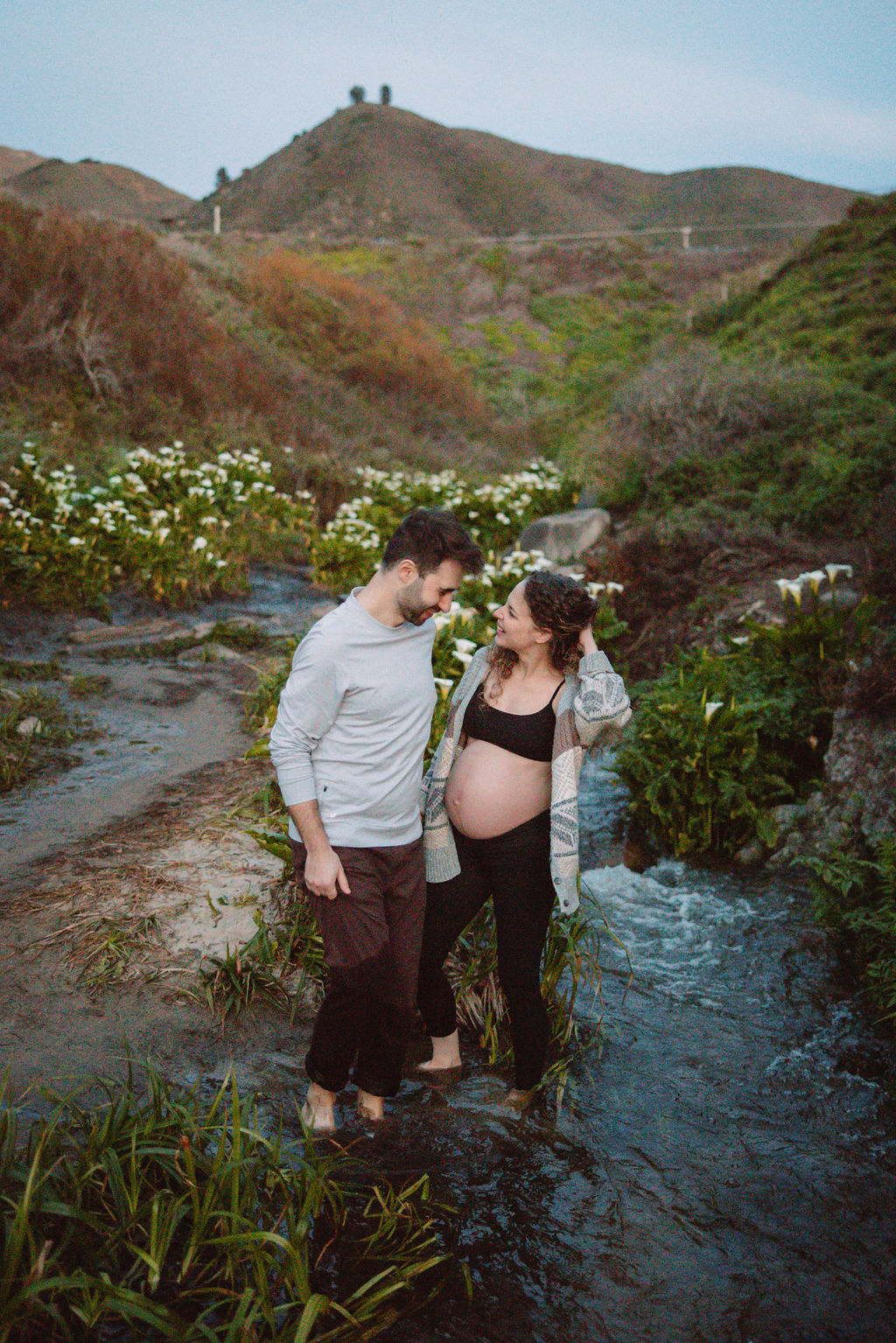 A couple expecting a baby, sharing a tender moment amidst a field of white flowers at Big Sur