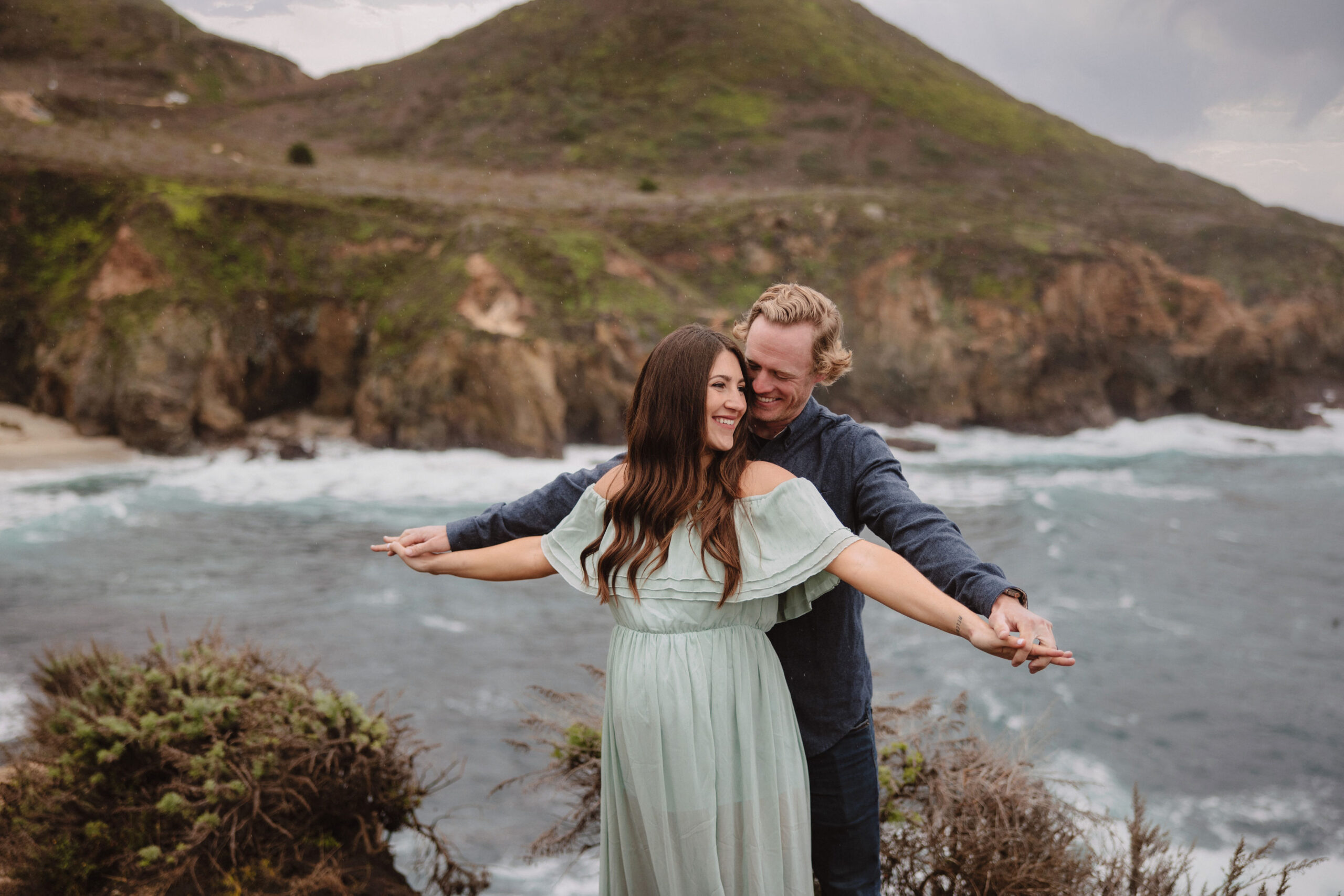 A couple embracing and smiling while standing on a coastal cliff with waves crashing against rocks in the background.