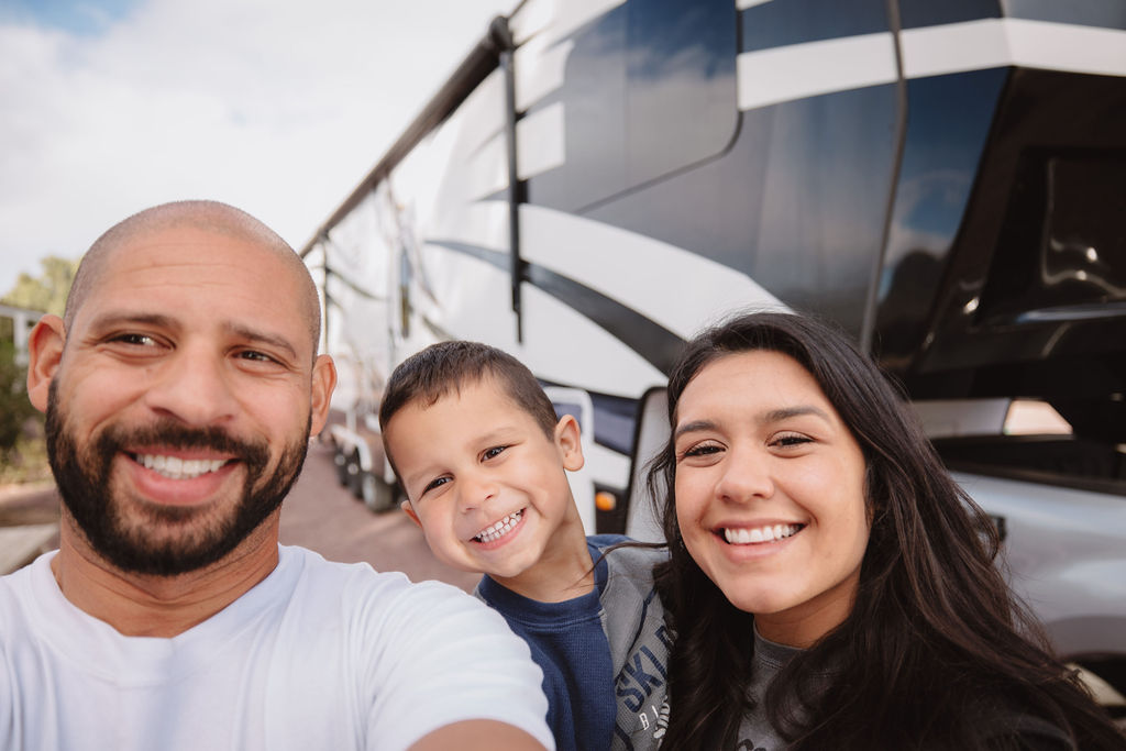 A family of three—two adults and a child—smiling for a selfie with an rv in the background on a sunny day.