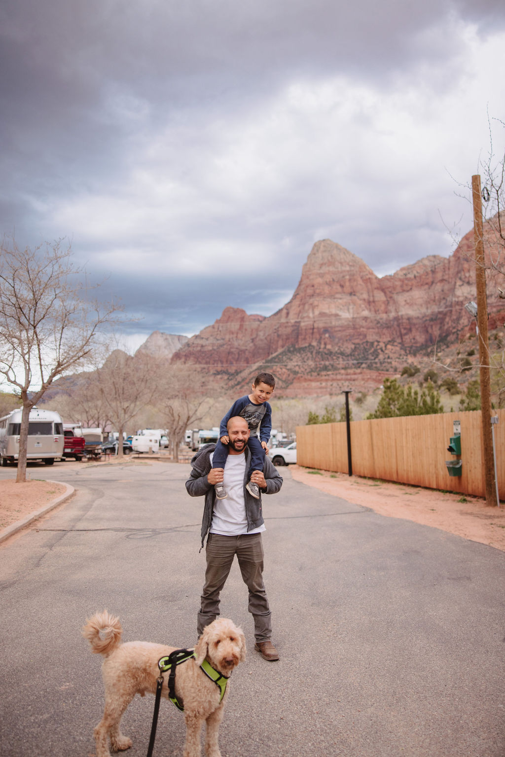 A man with a child on his shoulders and a dog on a leash walking on a road with red rock mountains in the background.