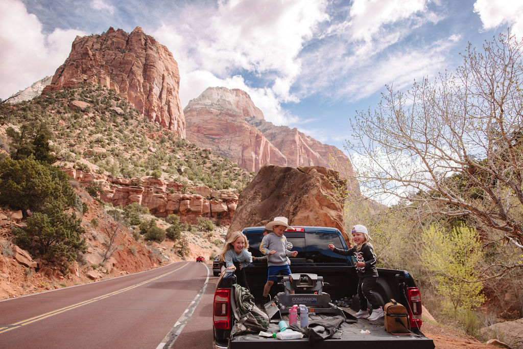Three people relaxing in a pickup truck's open trunk on a scenic road with red rocky cliffs and lush greenery in the background.
