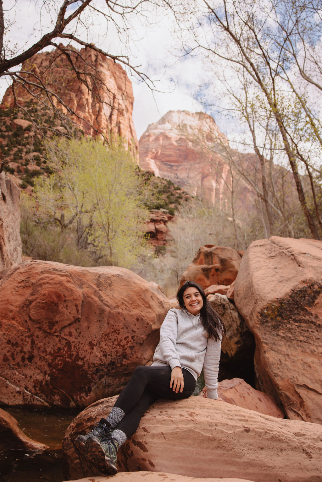 A woman sitting on a red rock beside a stream, smiling, with tall red cliffs and trees in the background at zion national park