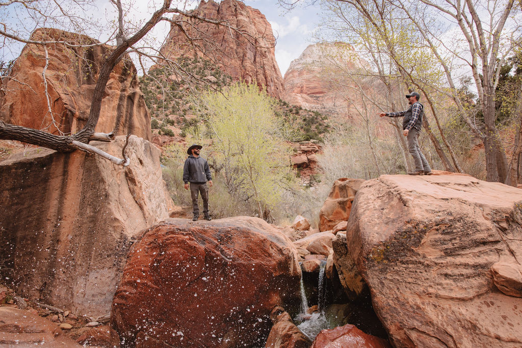 Two people exploring a rocky stream bed surrounded by red boulders and leafless trees in a canyon at zion national park