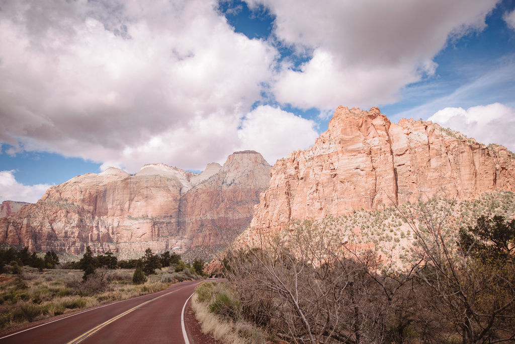 A curving road through zion national park with towering red rock cliffs and green foliage under a cloudy sky.