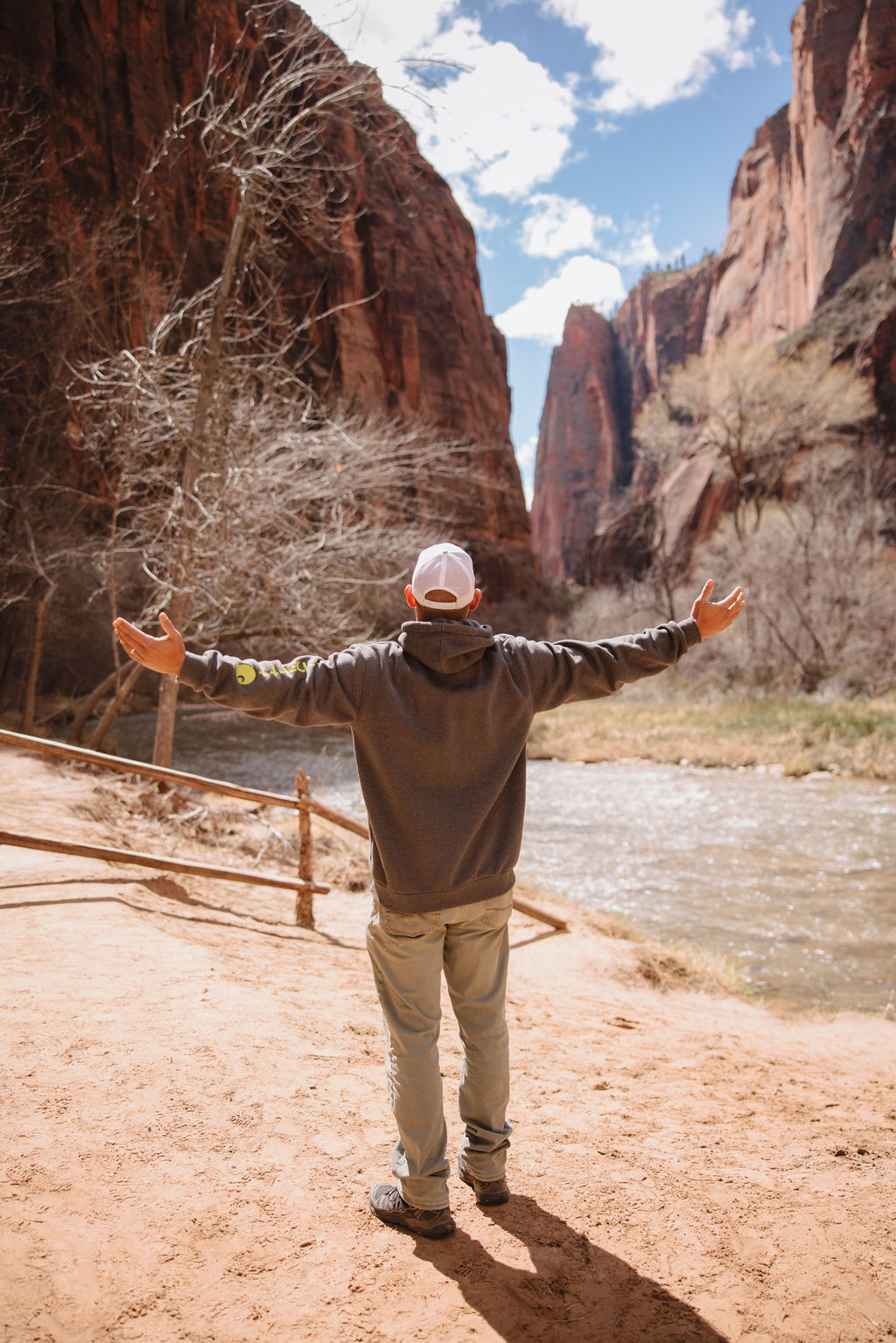 A man in a cap and jacket stands with arms outstretched facing a river and red rock canyon walls.