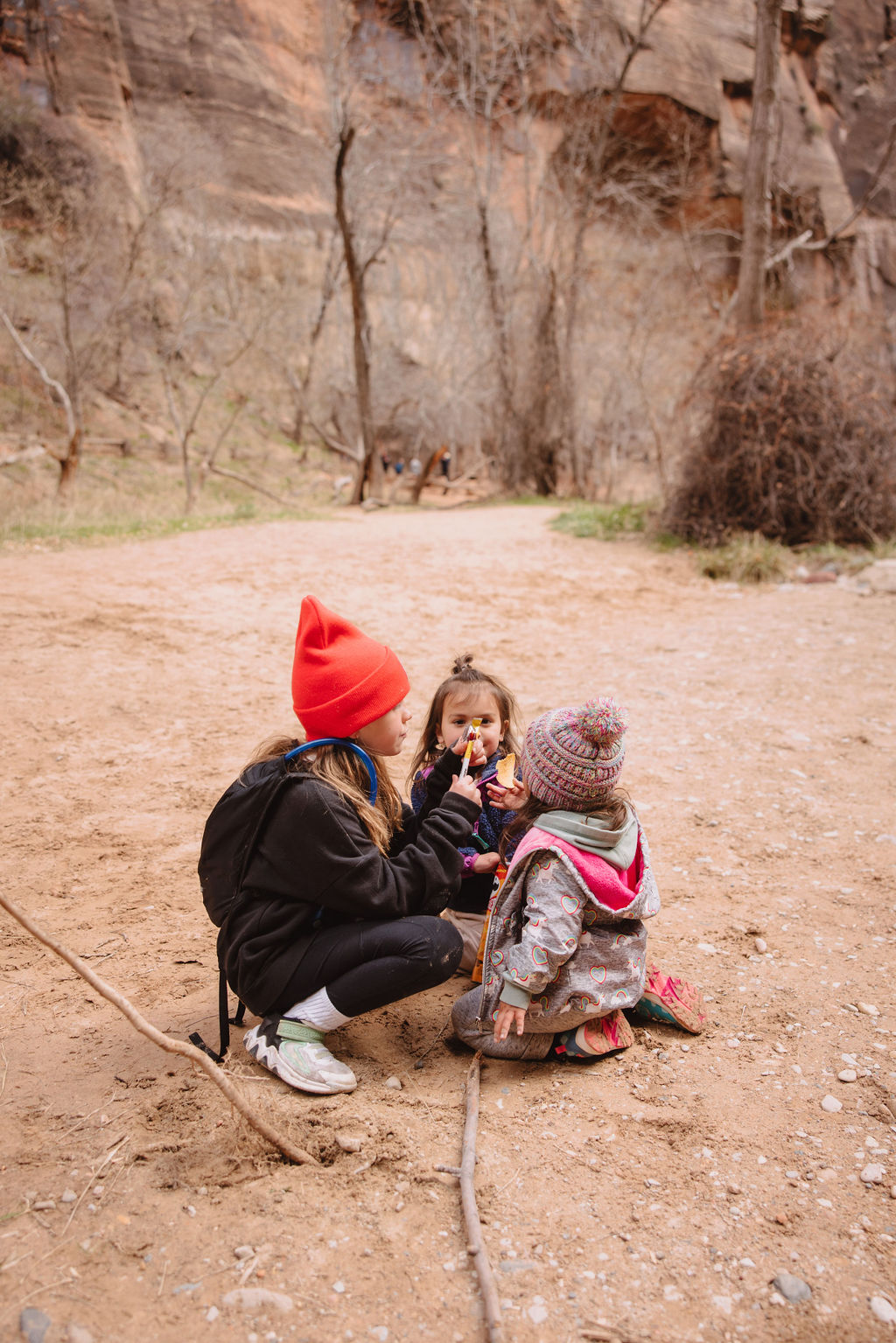Two young girls and a toddler play on a dirt path with a forested canyon background. one girl wears a red beanie, the other two are in colorful knit hats