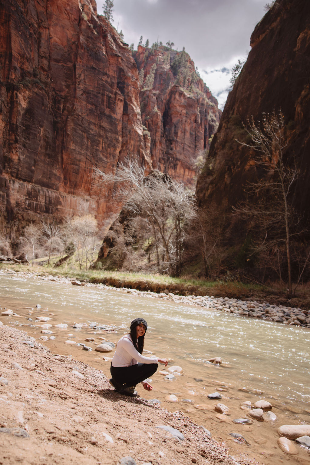 A person squats on a sandy riverbank, gazing at the camera, with a flowing river and tall, reddish cliff walls in the background at zion national park