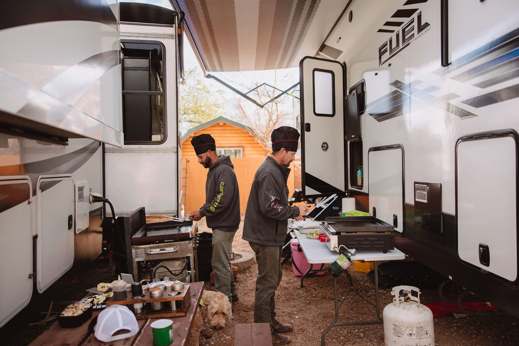 Two men cooking on an outdoor grill beside parked rvs in a wooded area. one is seasoning food while the other watches.