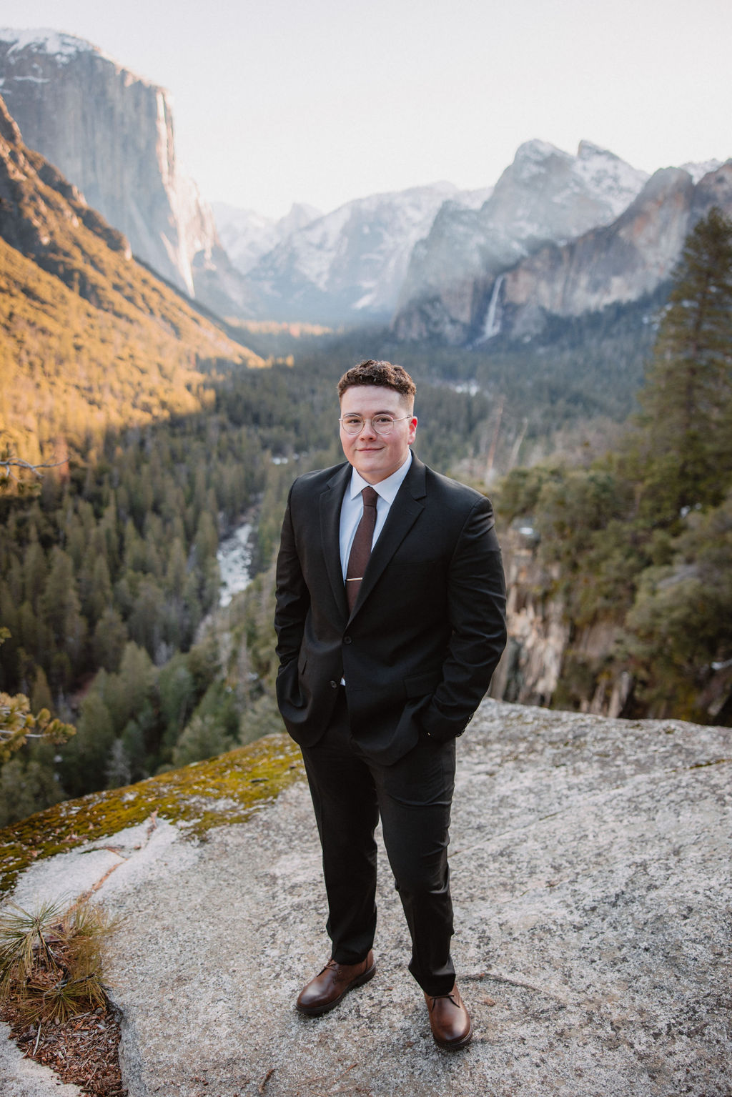 A groom in formal attire standing on a rocky overlook with a scenic mountainous background.
