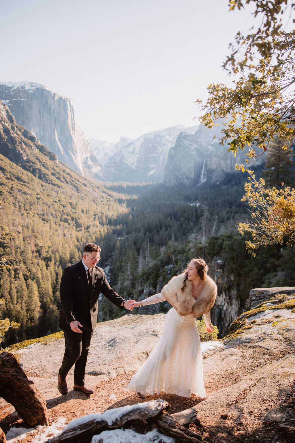 A bride and groom holding hands in a mountainous landscape, with the bride looking at the groom and the groom looking at the scenery.