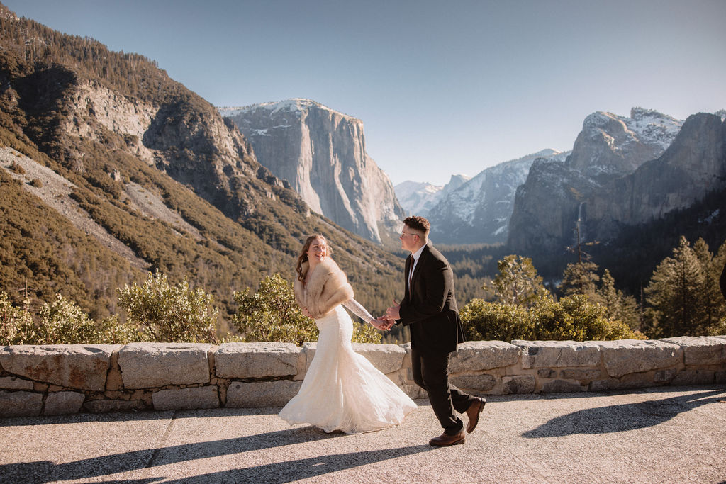 A couple in wedding attire holding hands, walking along a scenic overlook with mountains in the background.