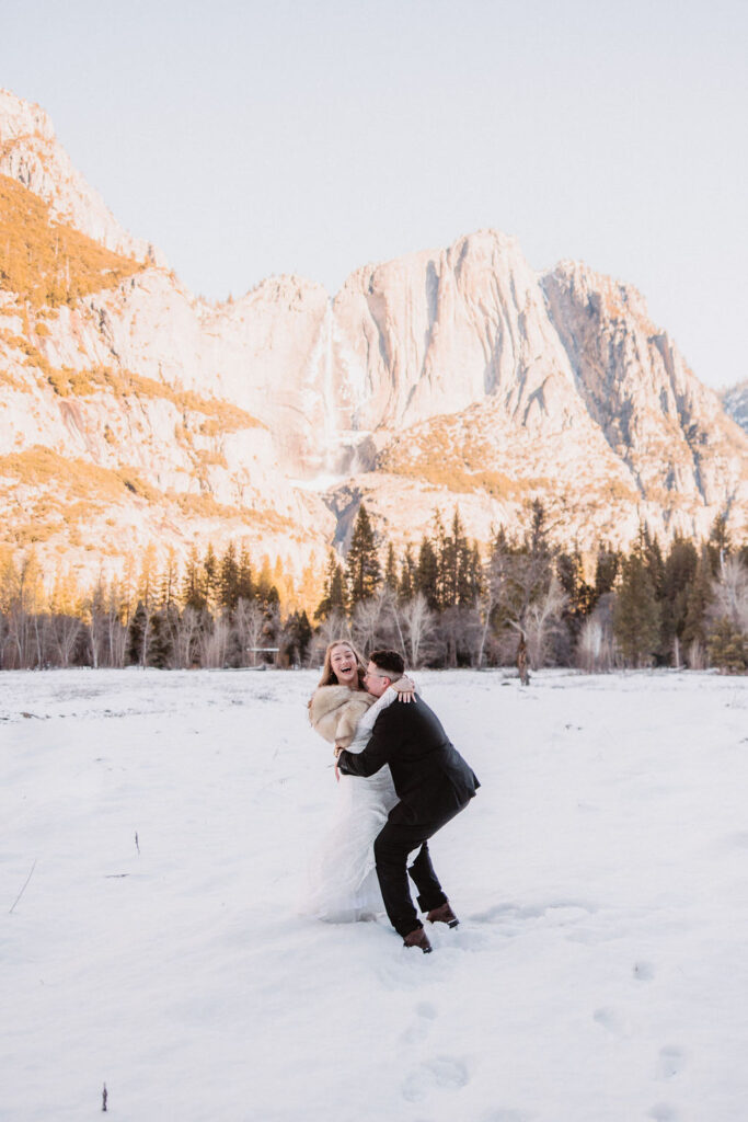 A couple in wedding attire sharing a playful moment in a snowy landscape with a mountain in the background.