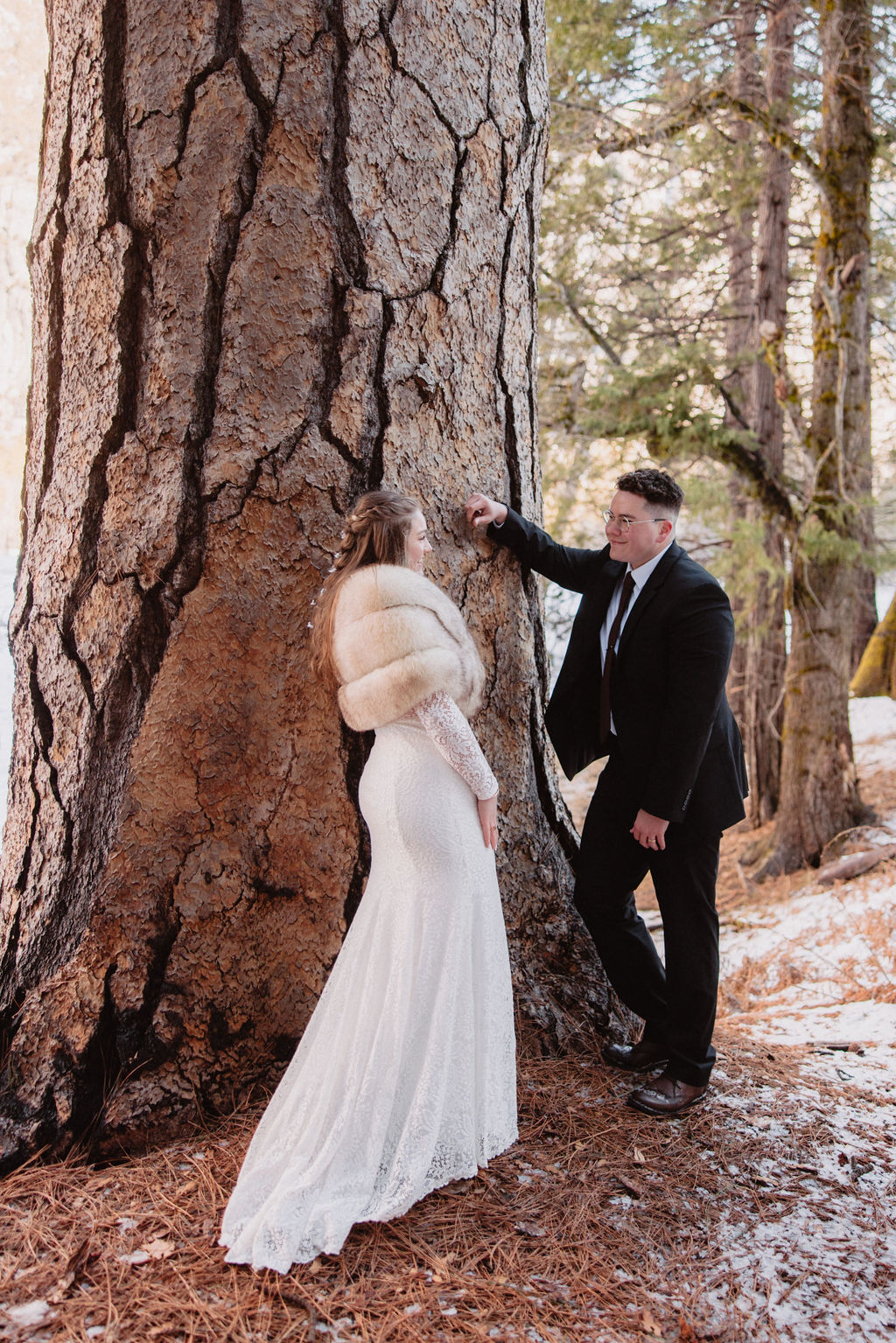 A couple dressed in wedding attire standing by a large tree in a snowy landscape at Yosemite National Park