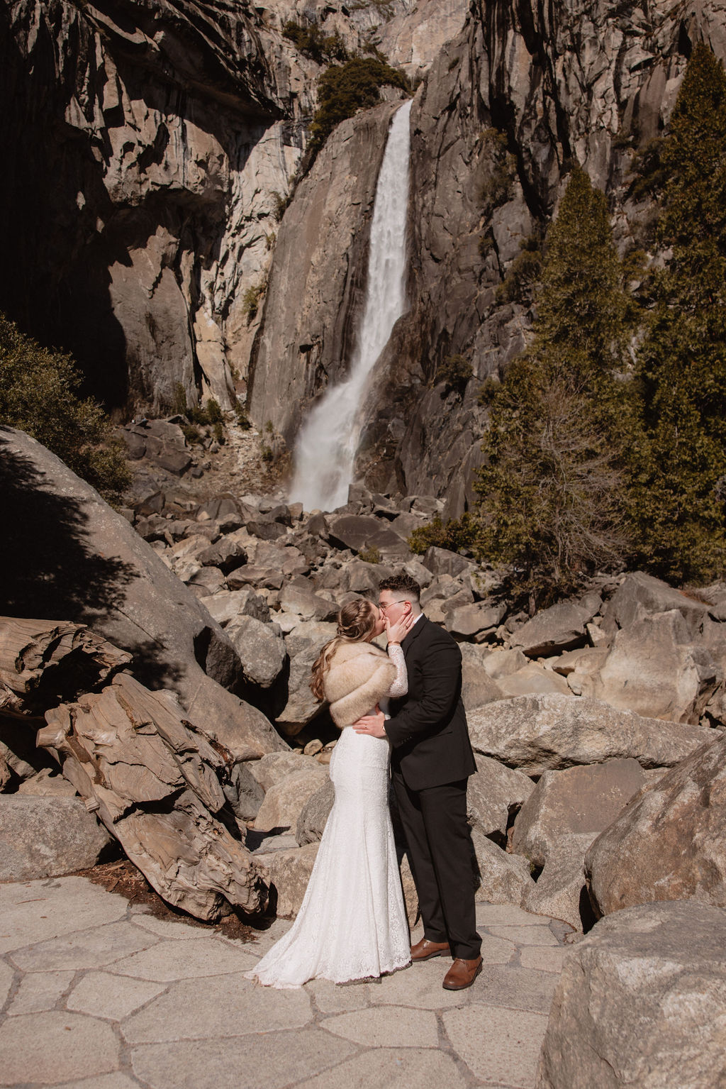 A couple kissing by a waterfall with rocky terrain around them.
