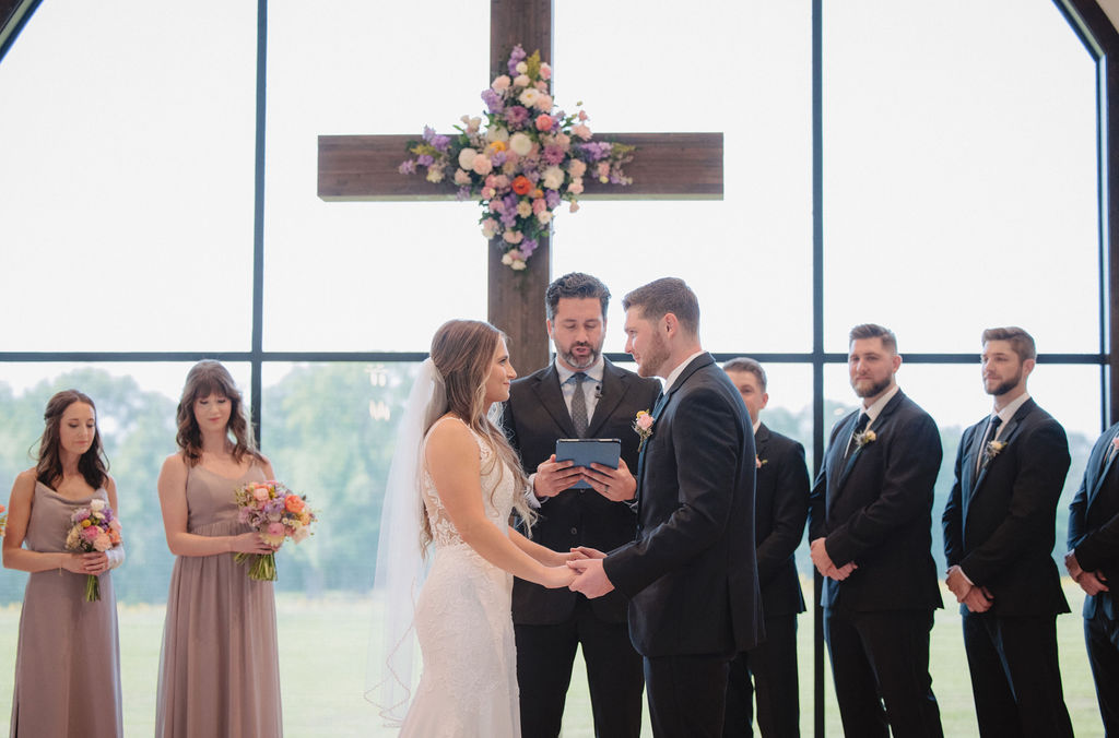A bride and groom stand facing each other, holding hands during their wedding ceremony. The officiant stands between them, holding a tablet. Bridesmaids and groomsmen are lined up on either side at deep in the heart farms