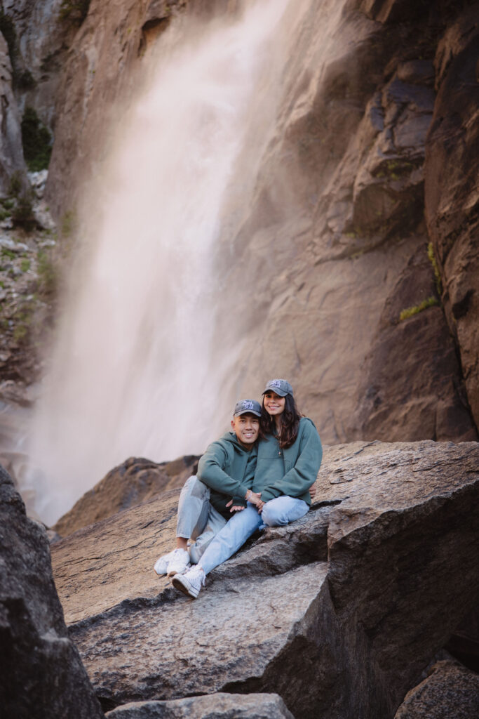 A couple stands close, touching their faces together, in front of a rocky landscape with a large waterfall called Yosemite falls in the background