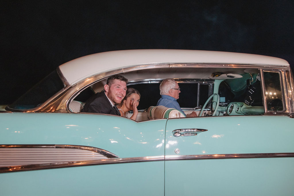 A man drives a vintage mint green car with a young couple smiling in the back seat at night after their wedding at deep in the heart farms