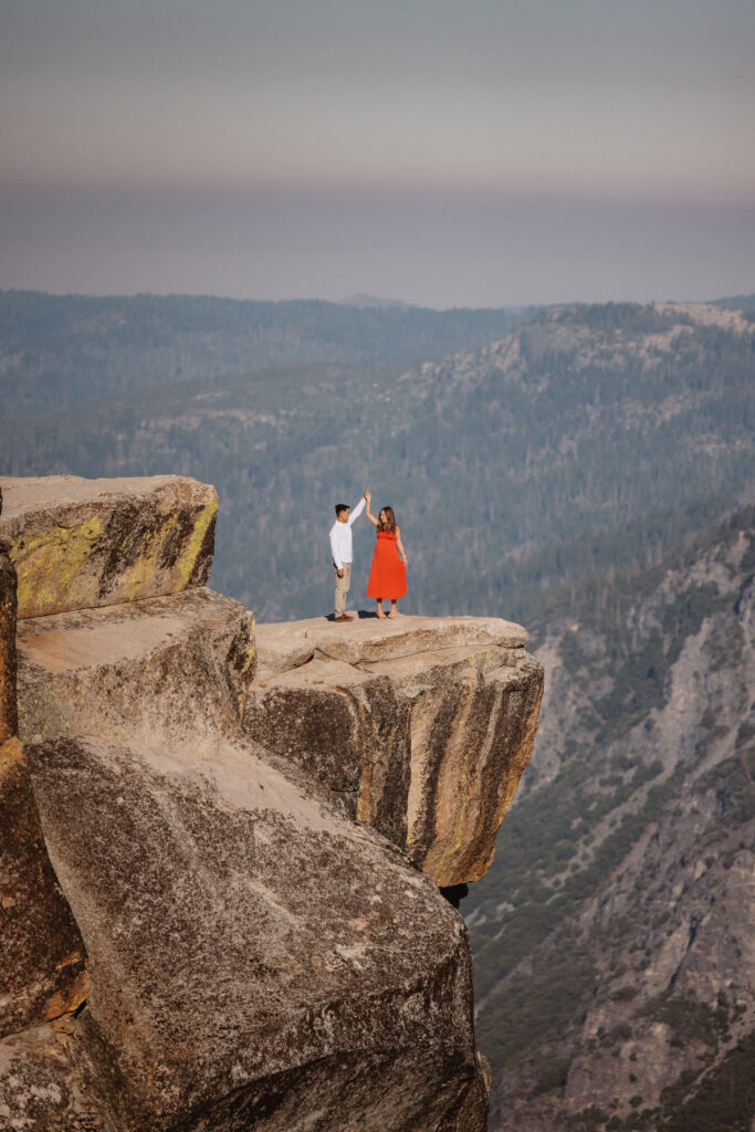 A couple stands on a rocky ledge with mountainous terrain in the background at Taft Point a Yosemite photo  location 
