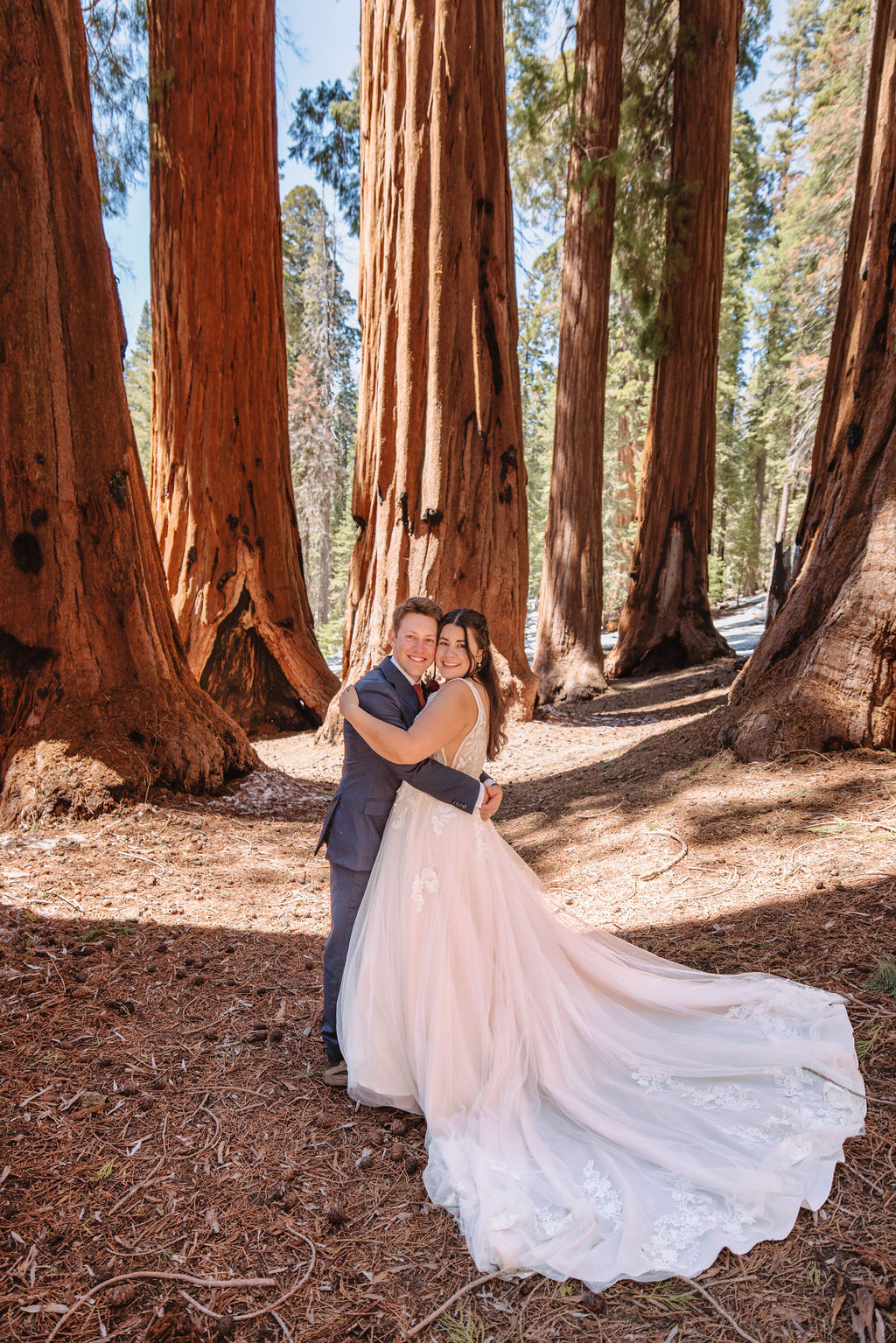 A bride and groom in a forest, surrounded by tall trees, with the bride wearing a long, flowing wedding gown at their Adventurous Elopement at Sequoia National Park