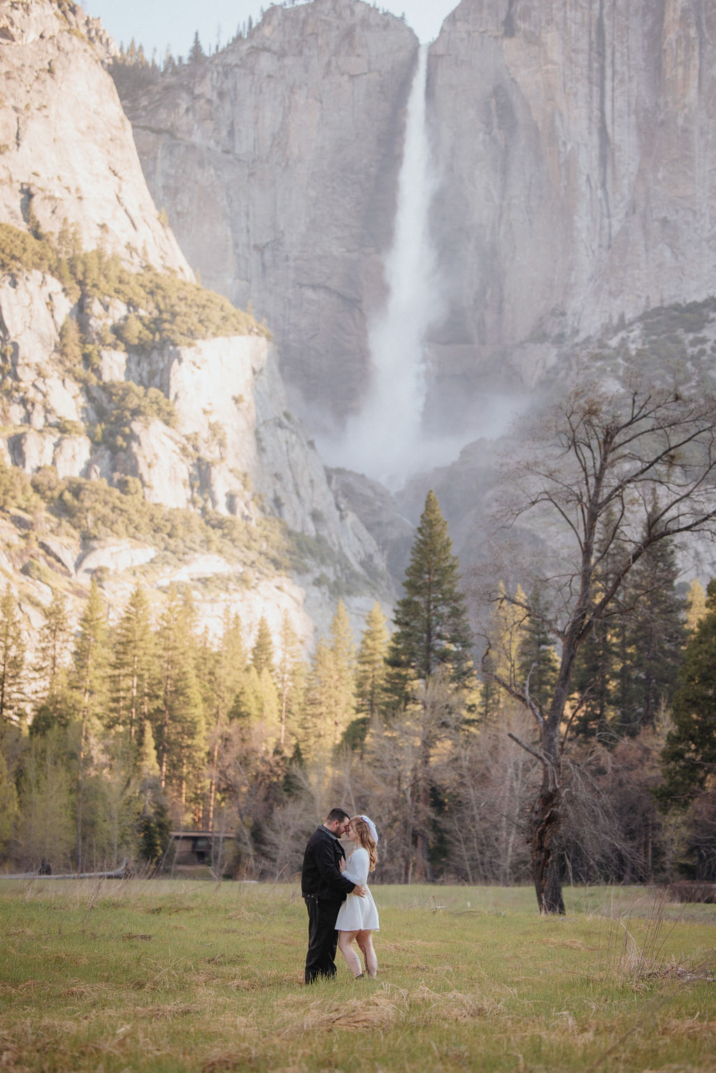 A man in a black hat and a woman in a white dress holding hands in a meadow, with tall misty cliffs in the background at their Yosemite engagement session