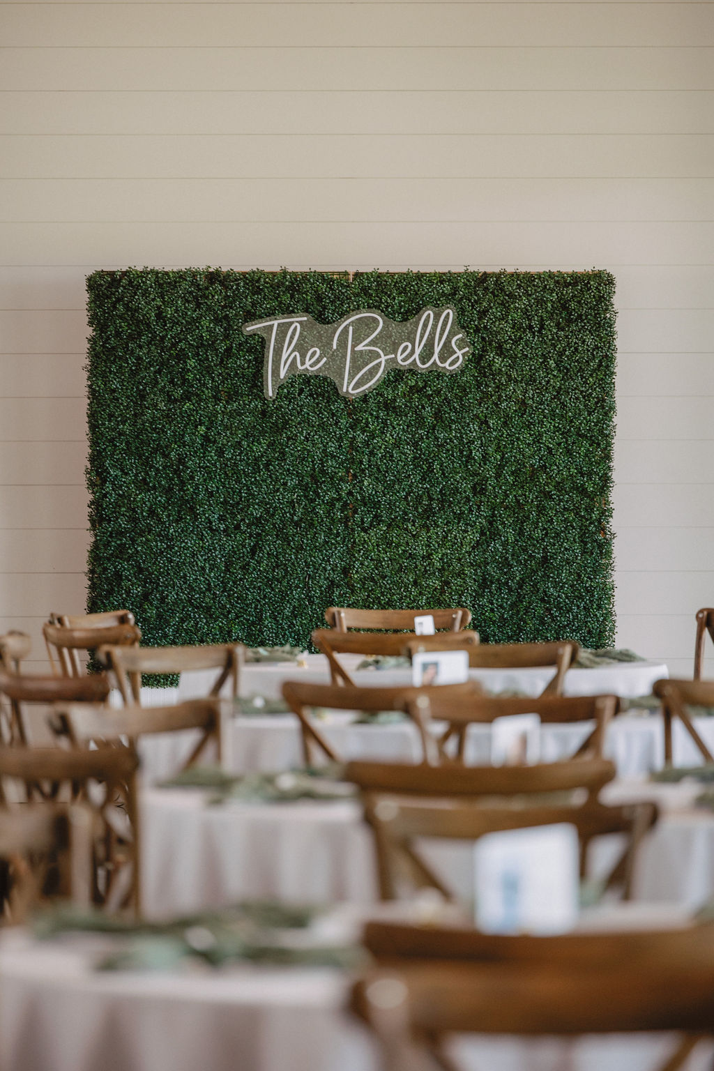 A green hedge wall decoration with the words "The Bells" in white script, positioned behind rows of wooden chairs and round tables covered with white cloths.