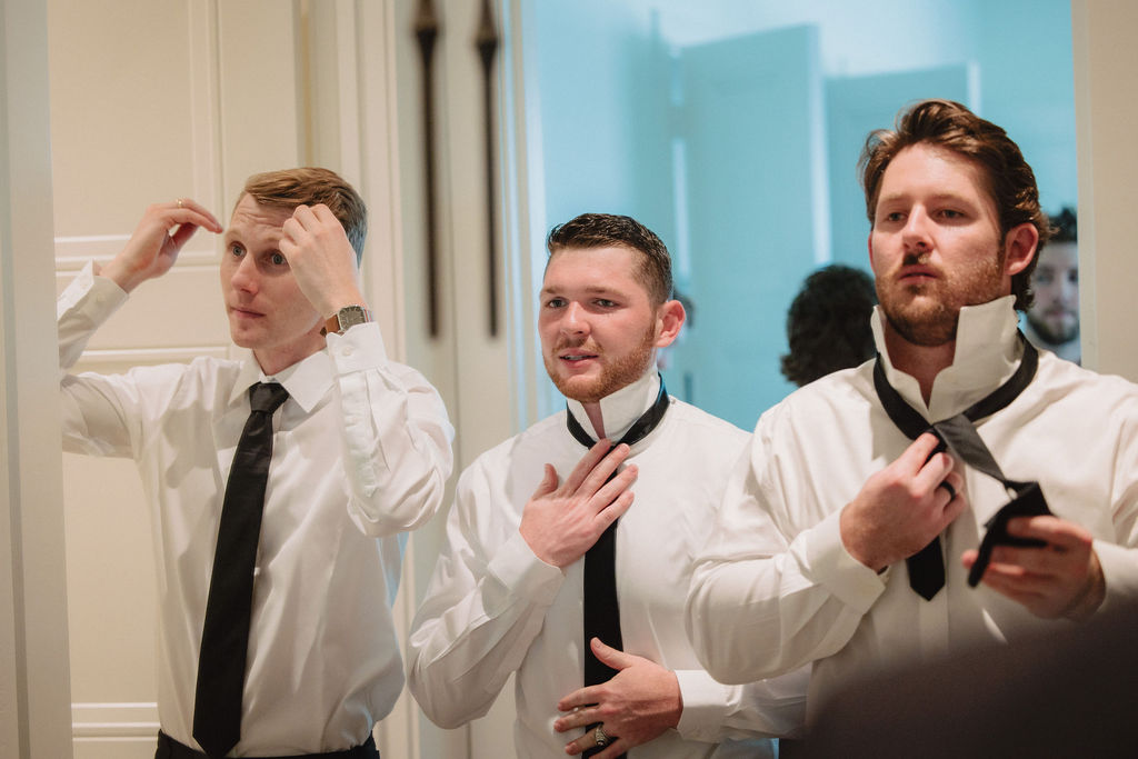 Three men in white dress shirts and black ties stand in front of a mirror adjusting their ties and fixing their hair.