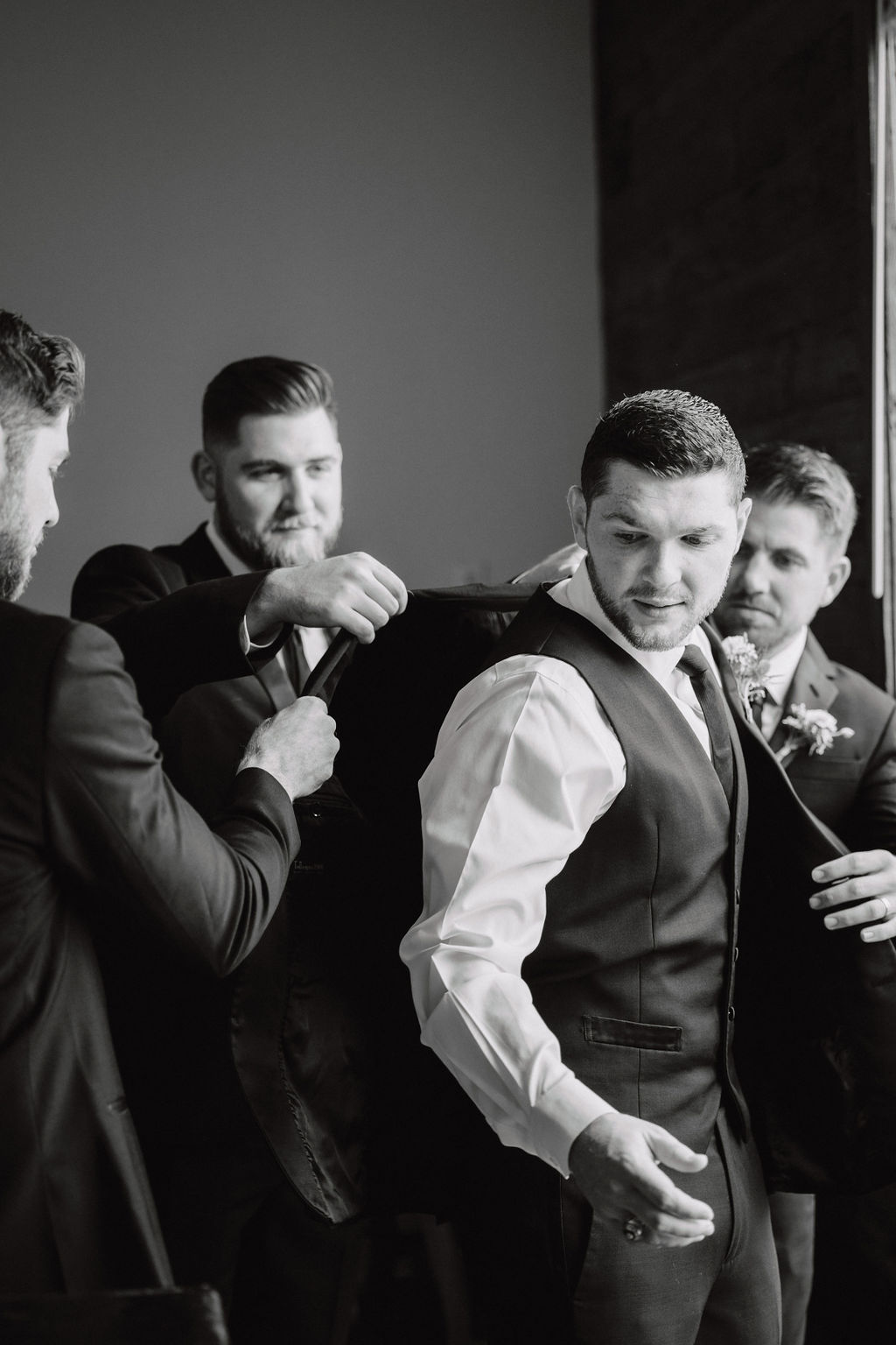 Four men, wearing formal suits, assist a groom in putting on his jacket. The atmosphere appears to be a preparation for a wedding
