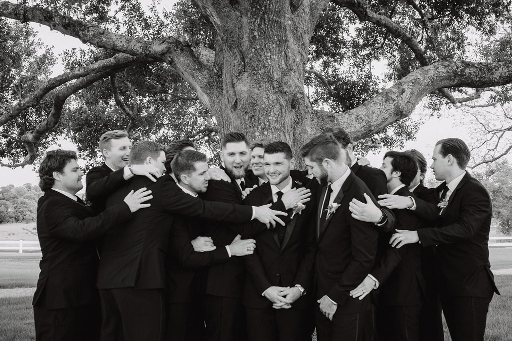 A group of men in suits are gathered under a large tree, some of them hugging the man in the center who is smiling at deep in the heart farms