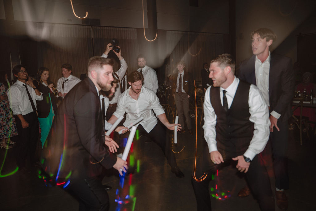 People at a wedding dancing energetically at an indoor event. Some are holding light sticks, and others are watching or taking photos.