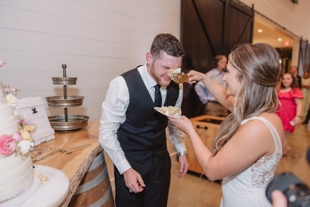 A bride playfully feeds cake to the groom, who is bending forward with a smile, while other people look on in the background.