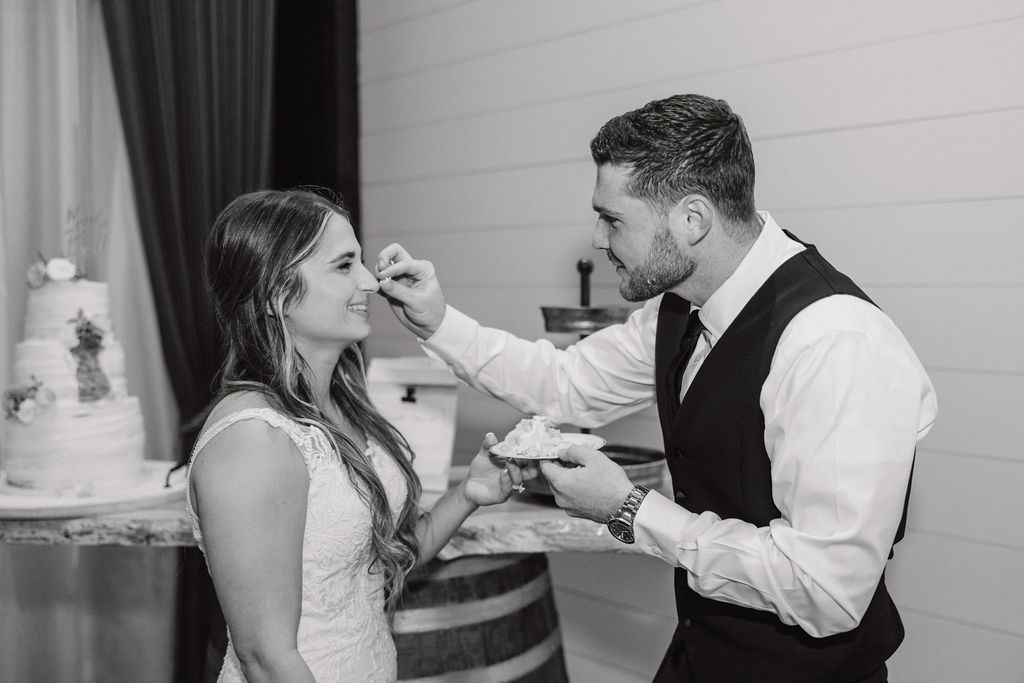 A bride playfully feeds cake to the groom, who is bending forward with a smile, while other people look on in the background.