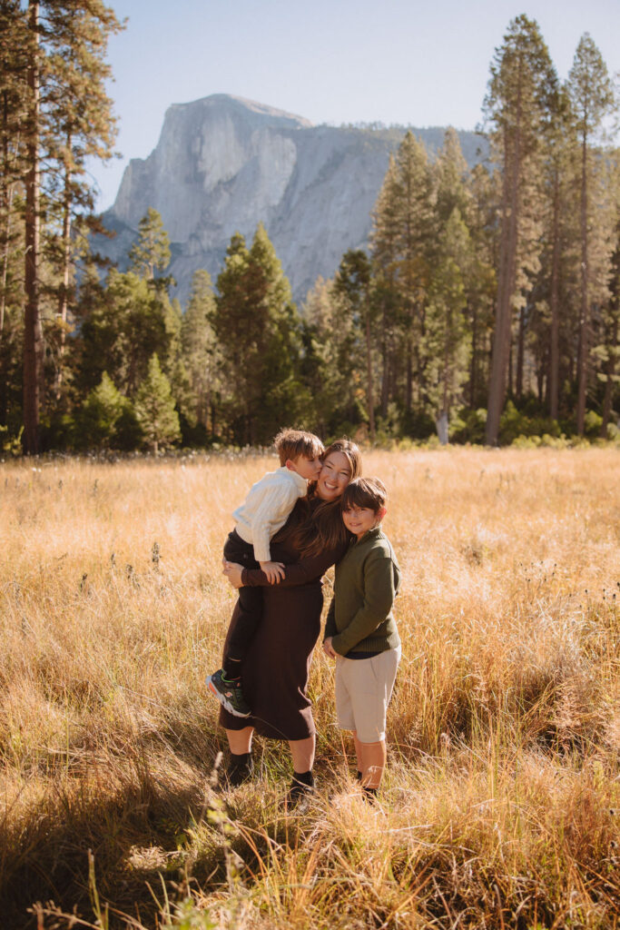 Two children laugh and play in a sunny, grassy field surrounded by tall pine trees, with a large rock formation visible in the background at Half dome meadow  in Yosemite  a Yosemite photo locations