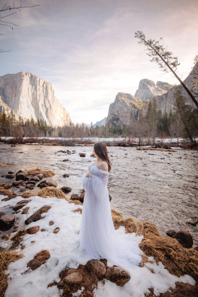 A woman dressed in a long white dress stands on a snowy riverbank, surrounded by tall mountains and trees, under a partly cloudy sky in Valley view in Yosemite photo locations