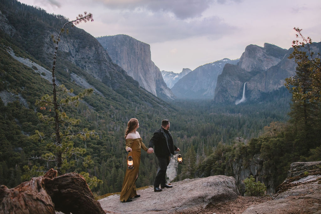 Couple embracing on a cliff with a scenic view of yosemite valley in the background. the woman wears a gold dress, and the man is in a dark suit at their yosemite engagement session